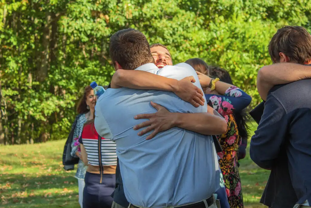 Two men embracing on a lawn, one laughing over the other's shoulder, with others hugging behind them
