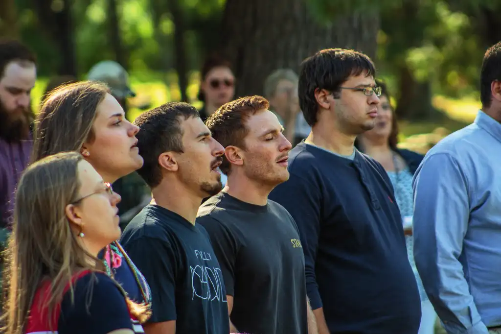 Men and women standing outdoors under trees, several singing with their eyes closed