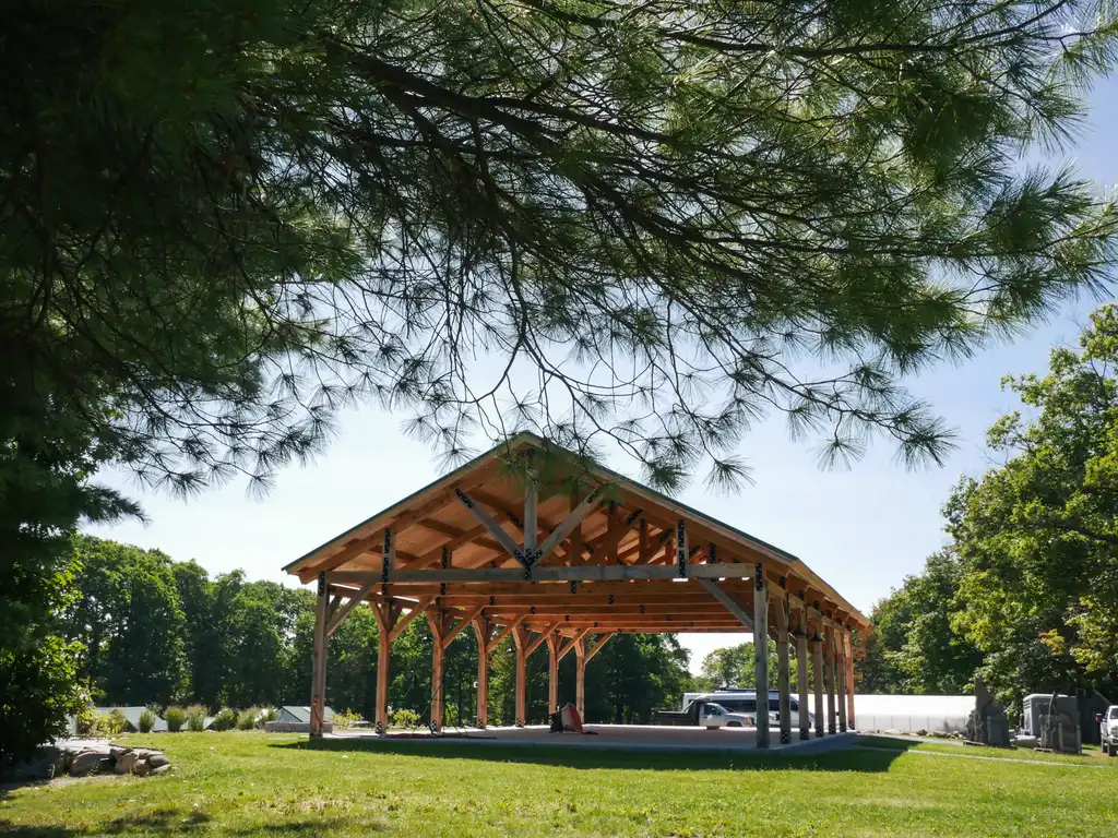 A large open timber-frame pavilion on a mown lawn, framed by pine boughs and green trees under a blue sky