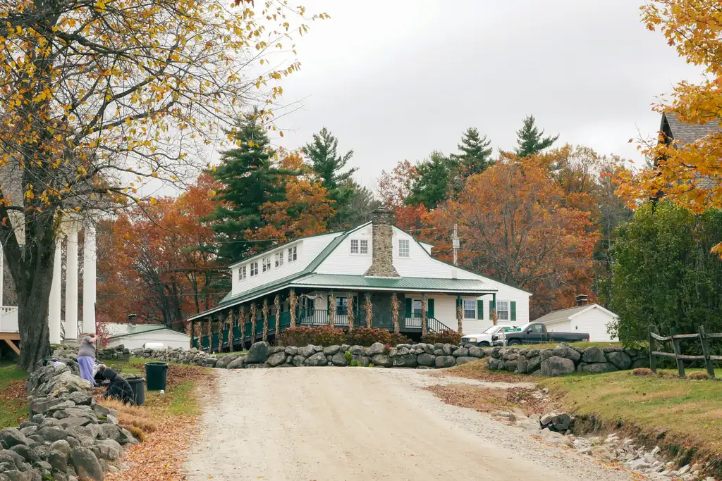 White building with a stone chimney and green wraparound porch at the end of a gravel road, framed by autumn maples