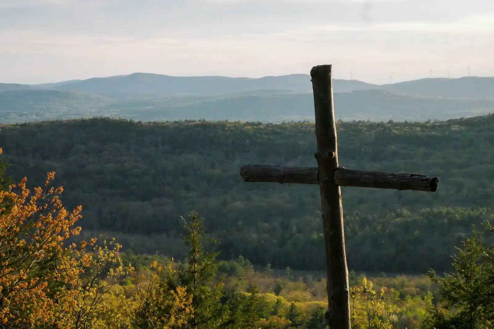 A rough wooden cross above orange and green treetops, looking out over a wooded valley to blue mountains