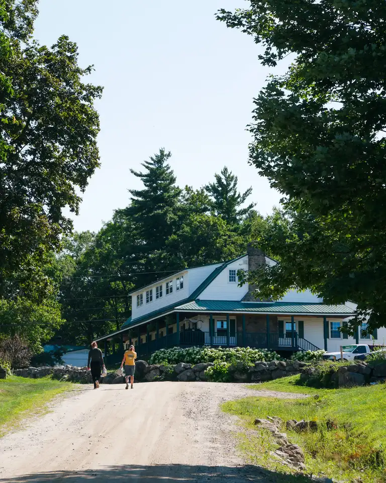 The same white house in summer under full green trees, two people walking up the dirt road toward it