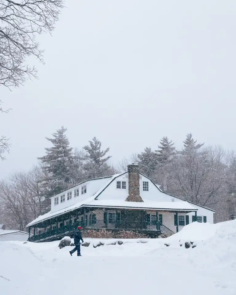 The same white house in winter, its roof and the yard covered in snow, one person walking across the yard