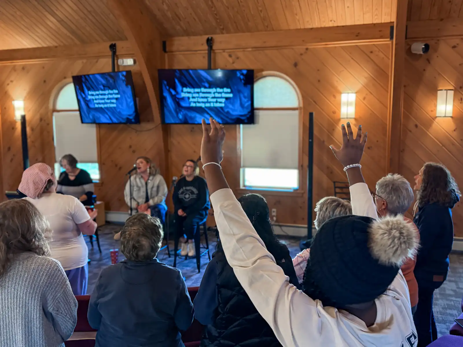 Women worship in a chapel, one with both hands raised, as three women lead singing beneath two lyric screens