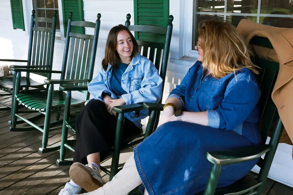 Two women in denim talk in green rocking chairs on a white clapboard porch with green shutters