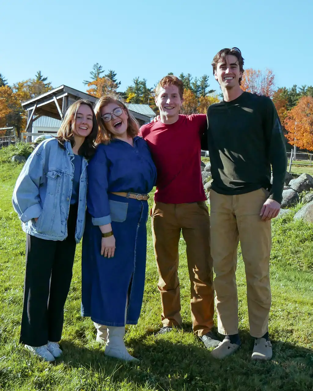 Four smiling young adults standing arm in arm on a sunny lawn, an open timber shed and autumn trees behind