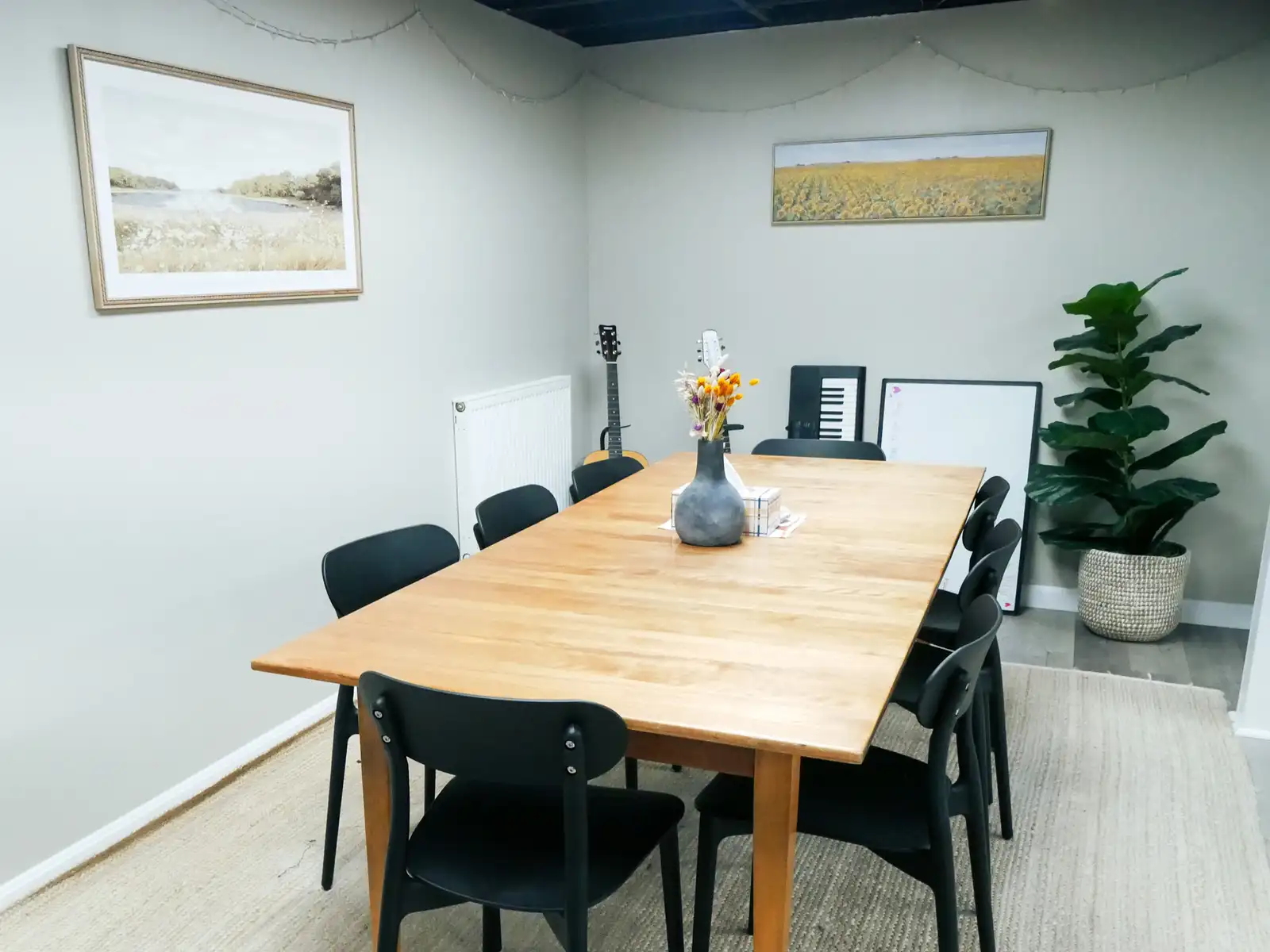 A long wooden table with black chairs in a bright room, with framed landscape prints and a tall potted plant