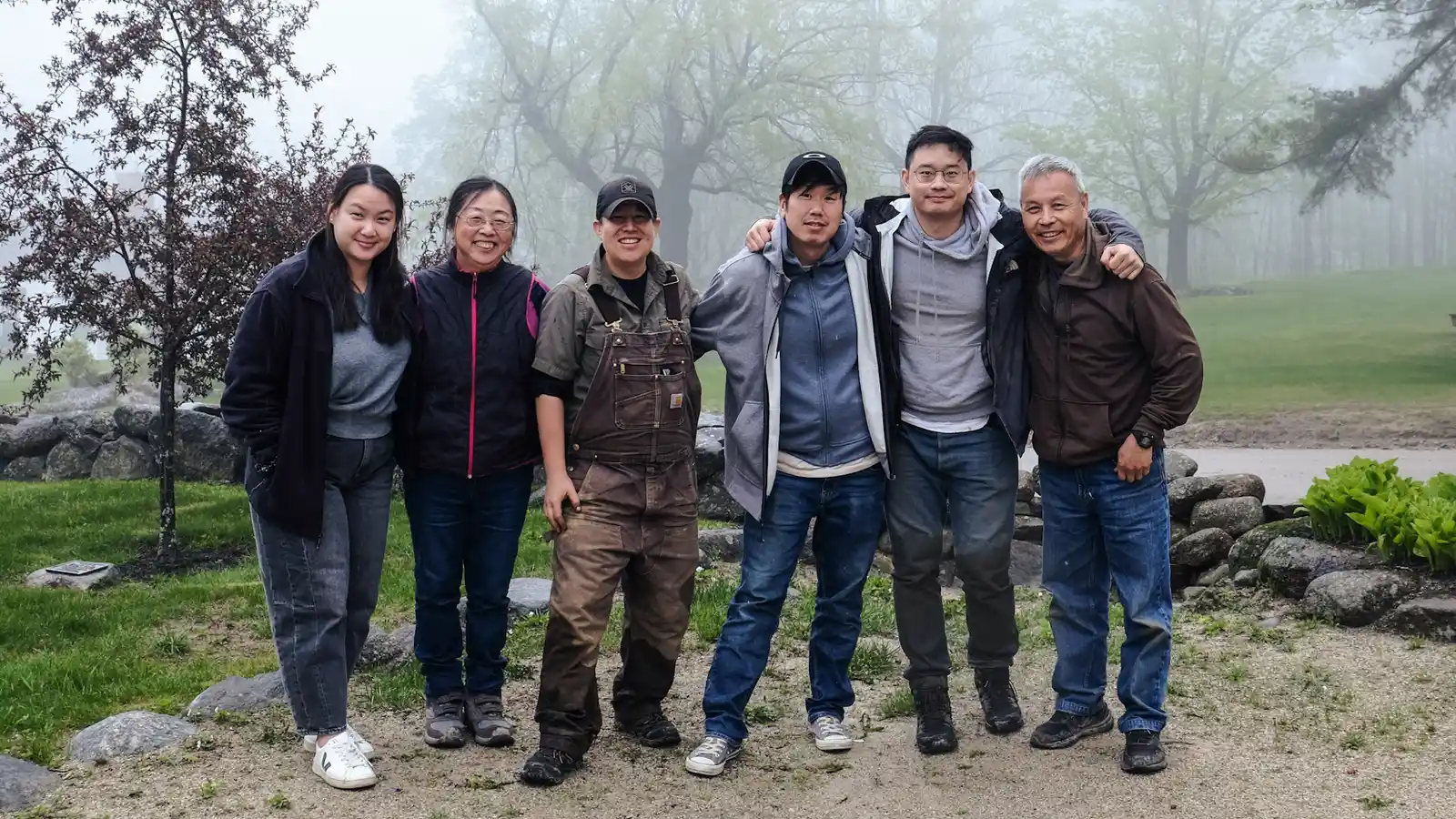 Six Work Group volunteers standing arm in arm on a gravel path by a stone wall on a foggy spring morning